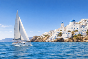 White sailing yacht cruising past a sunlit Greek island with whitewashed buildings and blue-domed churches above clear Aegean water.