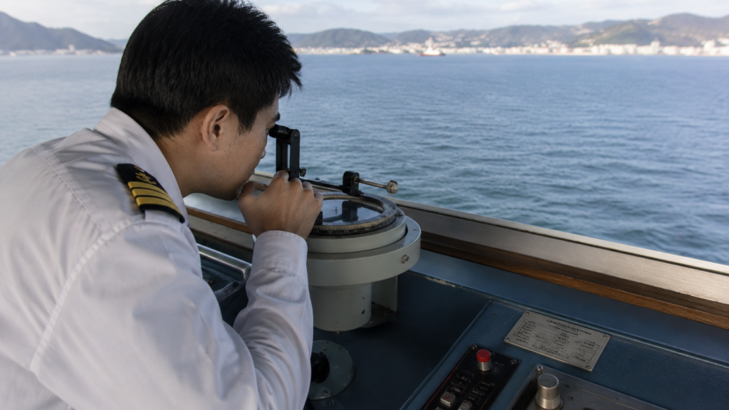 A navigating officer taking a compass bearing with an azimuth ring on the bridge wing, the coastline visible in the distance.